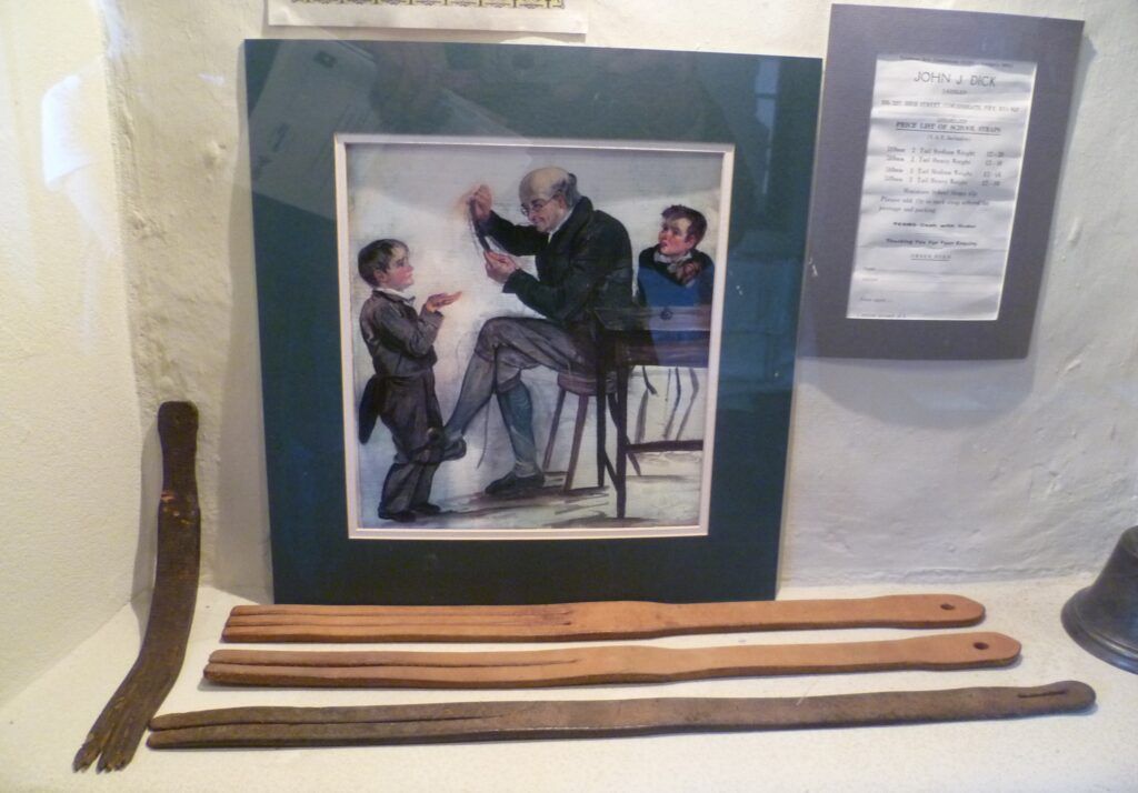 Museum display of leather tawses made in Lochgelly beside a John J. Dick price list and a painting of a schoolmaster striking a boys palm